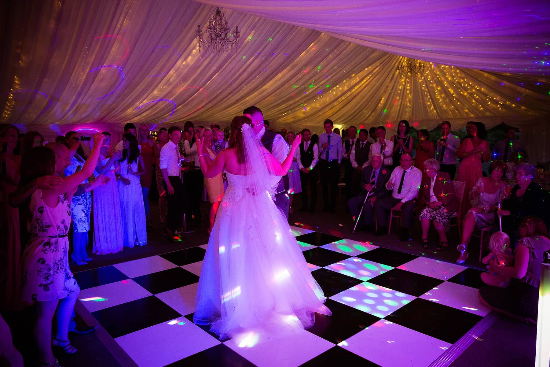 A bride in the middle of a lit chequerboard dance floor in a marquee, guests dancing all around her